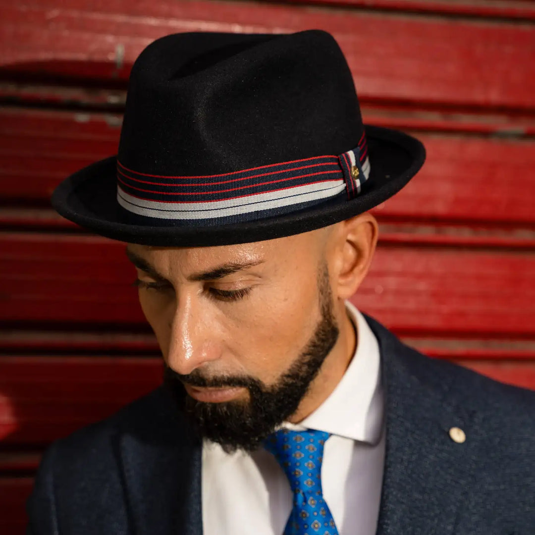 modello adulto con cappello lobbia homburg in feltro raso di lapin blu primario nesti completo blu navy e cravatta azzurra posa su sfondo rosso ritratto di profilo