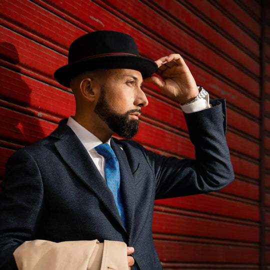 modello adulto con cappello lobbia homburg in feltro raso di lapin primario nesti e giacca in mano beige posa contro parete rossa look business