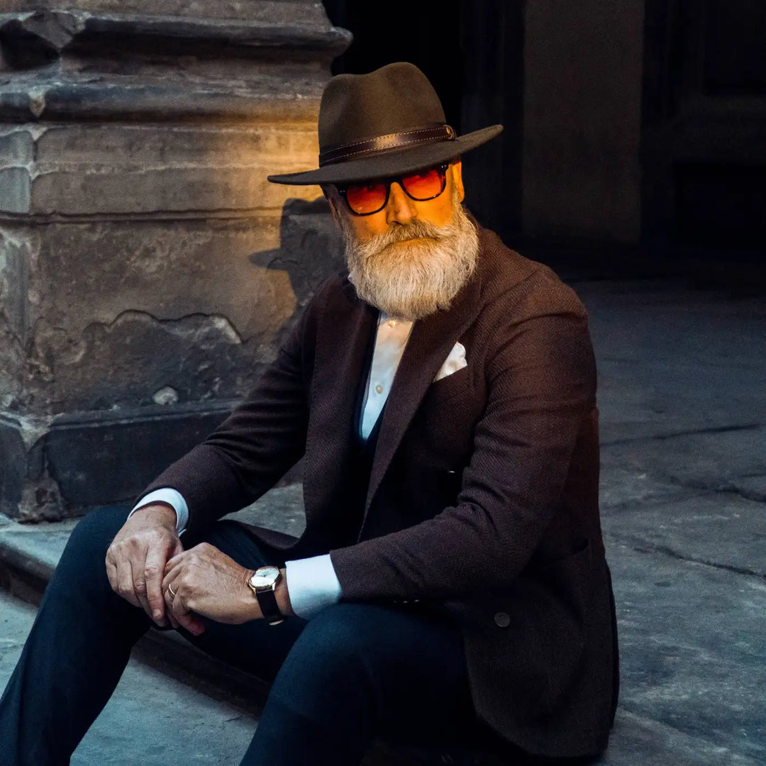 modello maturo con barba curato indossa cappello fedora tradizionale primario nesti antipioggia colore marrone con outfit scuro e ambientazione suggestiva serale