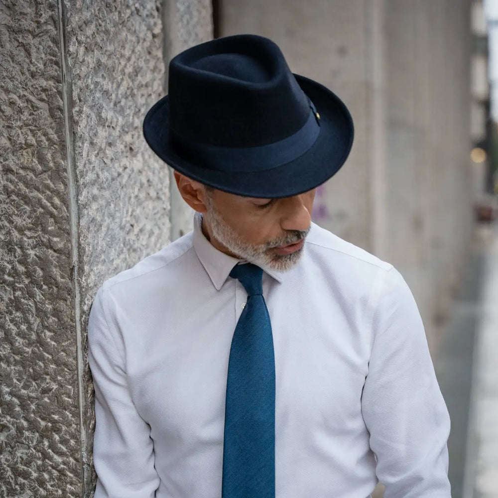 uomo elegante con trilby classico primario nesti lapin blu navy cravatta azzurra su camicia bianca posa raffinata contro colonna